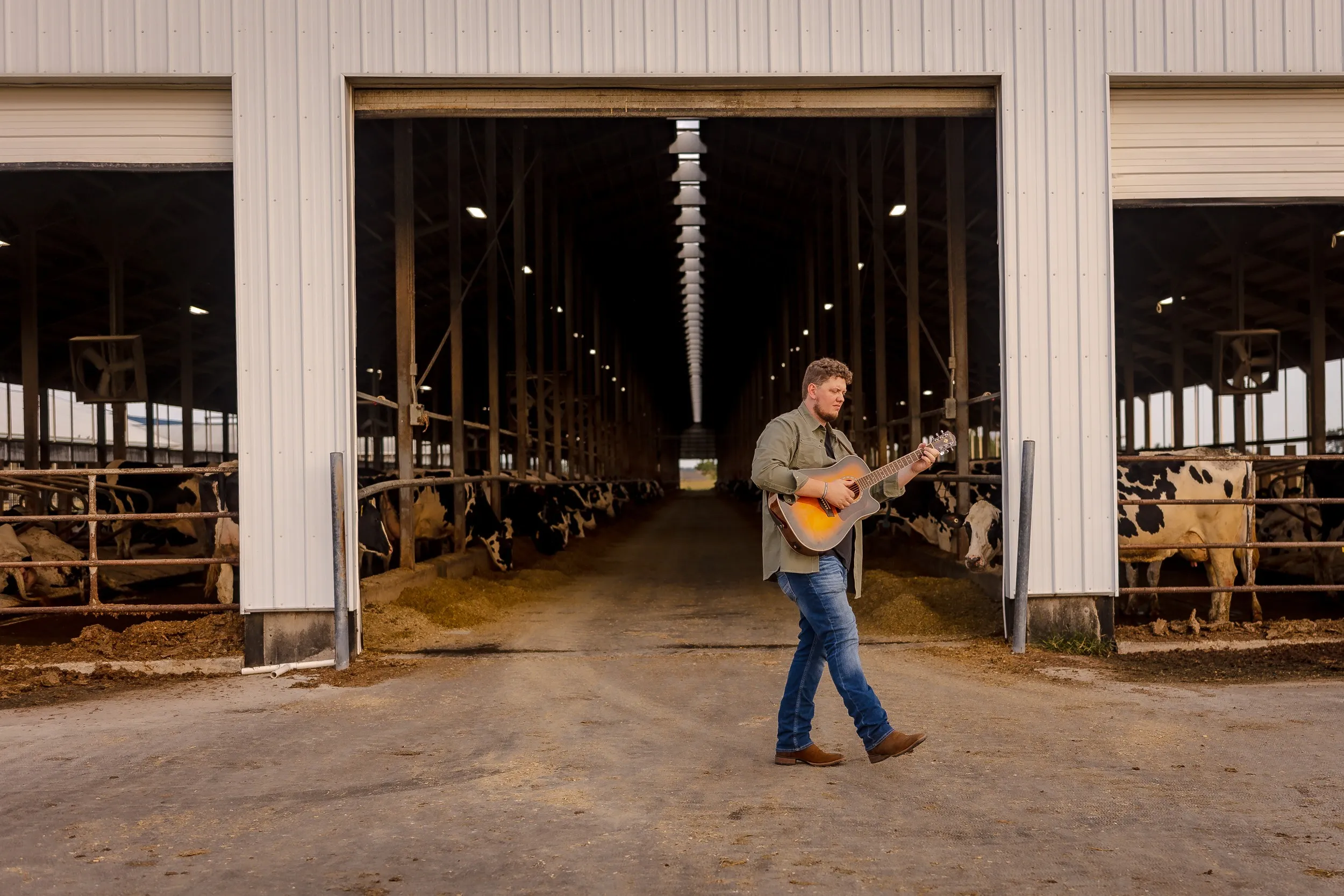 Isaac Buschur playing guitar as he walks past an open dairy barn