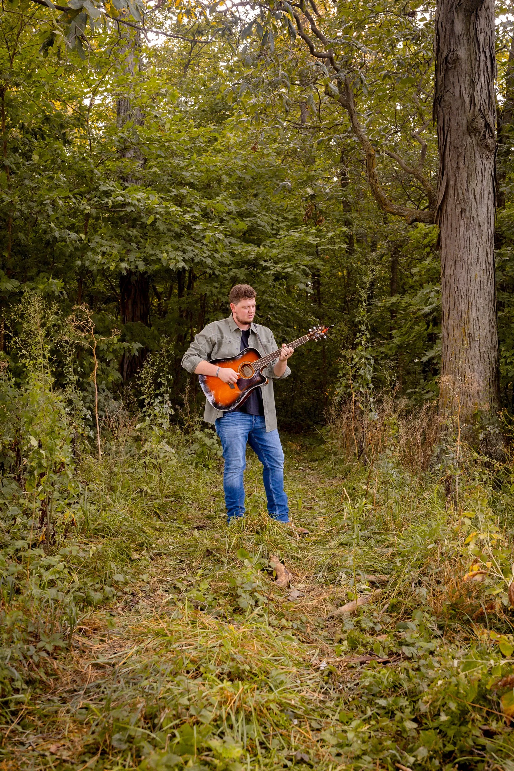 Isaac Buschur playing acoustic guitar on a wooded trail